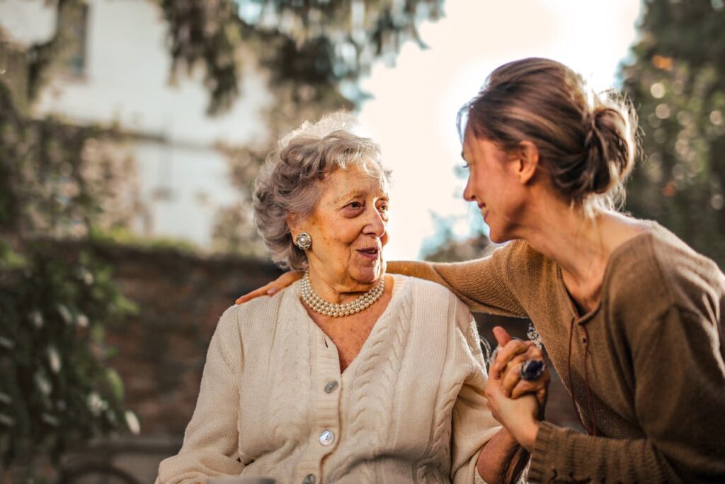 Eine Frau umarmt und hält einer Seniorin die Hand. Beide Personen lächeln.